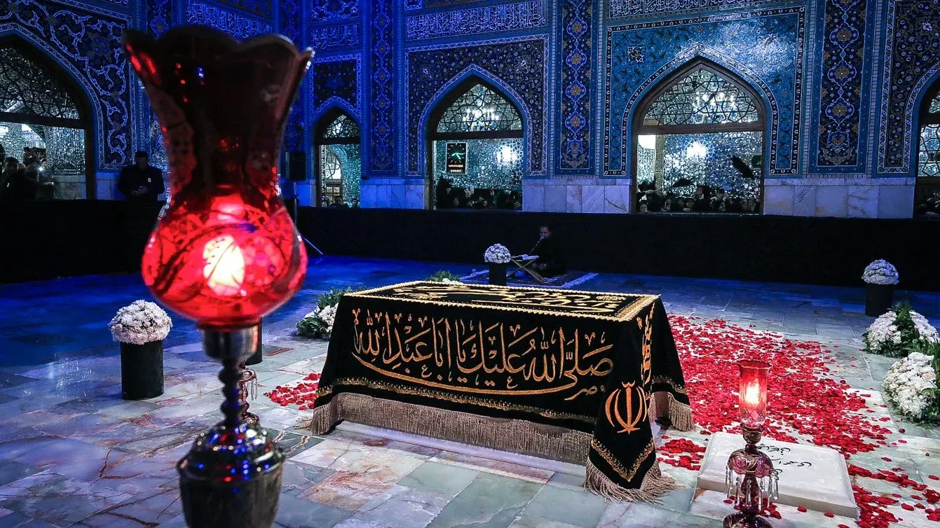 ضريح علي خامنئي في مشهد مغطى بغطاء أسود مزخرف وزهور بيضاء وحمراء، مع إضاءة خافتة تعكس أجواء الحداد والذكرى.