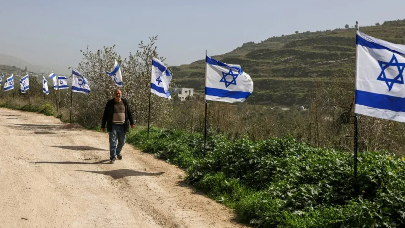 رجل يسير على طريق ترابي محاط بأعلام إسرائيلية في الضفة الغربية، مما يعكس التوترات السياسية حول الأراضي المحتلة.