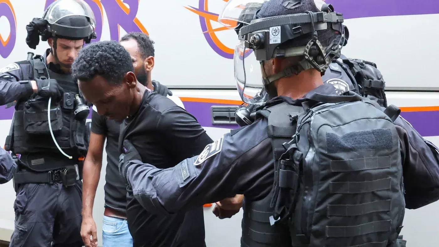 اعتقال طالب لجوء إثيوبي في إسرائيل أثناء تنفيذ الشرطة عملية ترحيل، مع وجود عناصر أمنية في الخلفية.