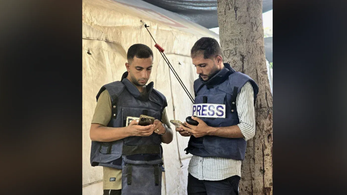 صحفيان يرتديان سترات تحمل علامة "صحافة" يتفقدان هواتفهما في موقع هجوم إسرائيلي على غزة، حيث قُتل زملاؤهم.