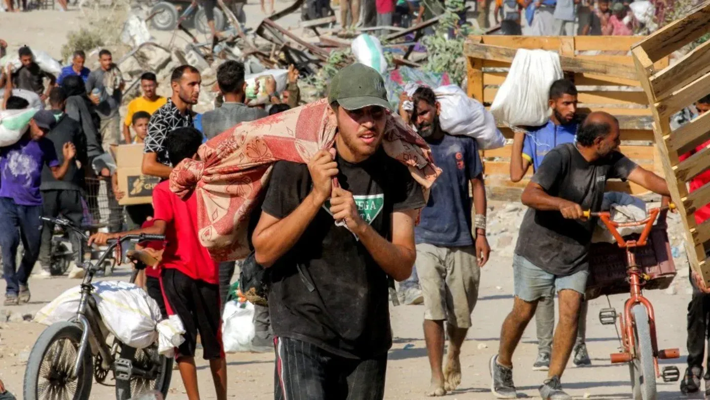 شبان فلسطينيون يحملون مساعدات إنسانية تحت نيران كثيفة في منطقة زيكيم، مع وجود حطام خلفهم، مما يعكس الوضع الصعب في غزة.