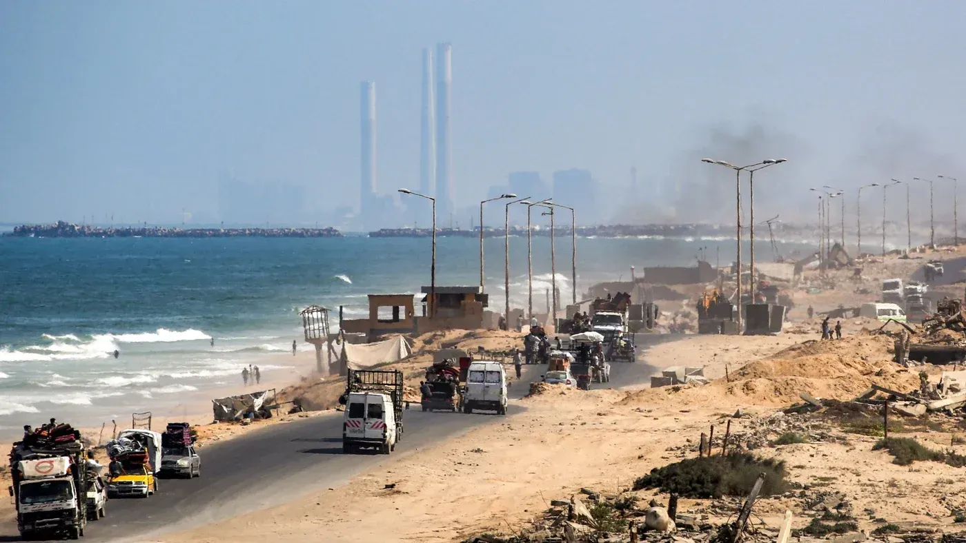 صورة تظهر الطريق الساحلي في غزة مع شاحنات وأعمال إعادة الإعمار، خلفها البحر وأبراج كهرباء، تعكس جهود التنمية الاقتصادية في المنطقة.