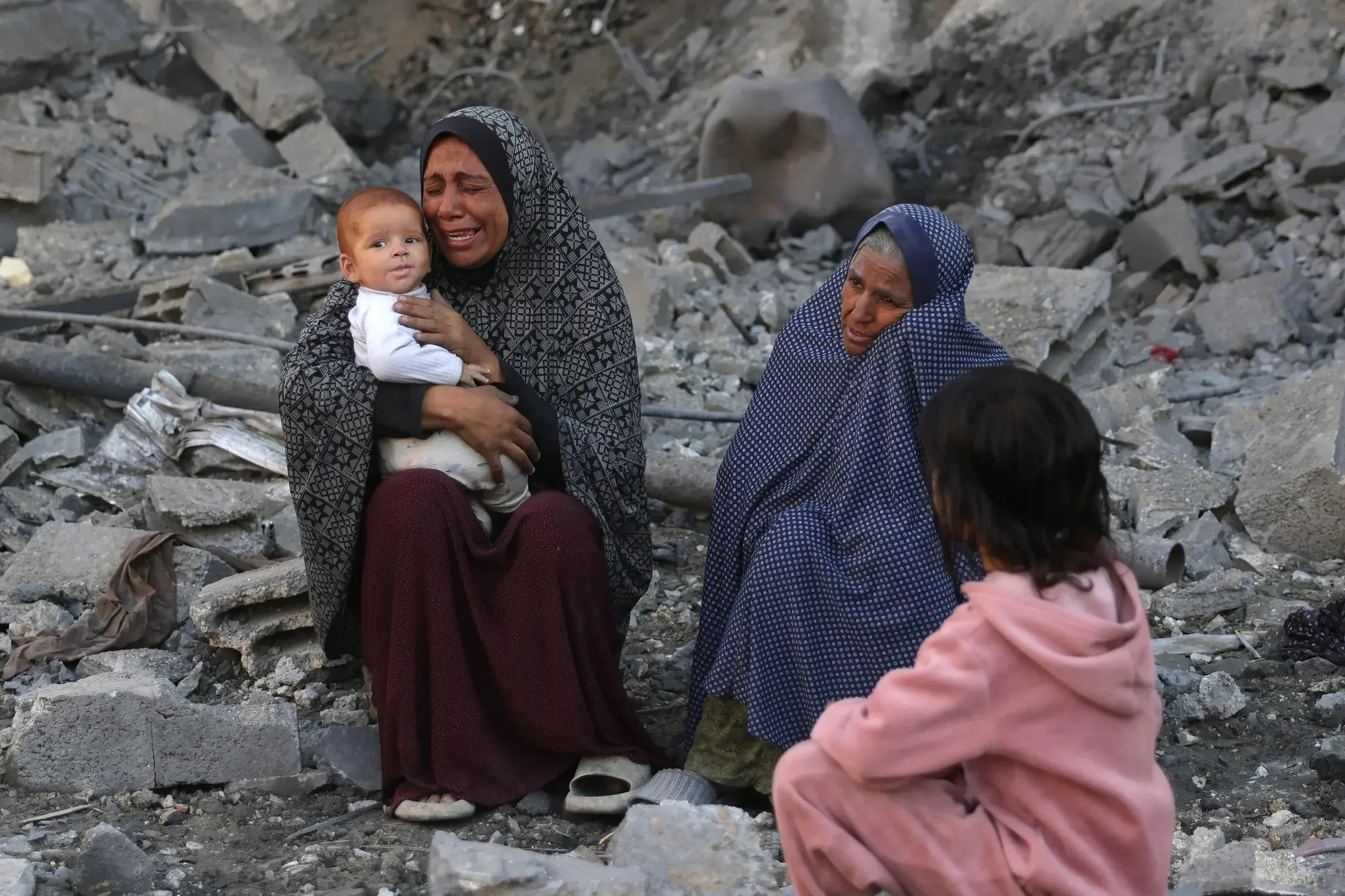 نساء وأطفال يجلسون على أنقاض مبنى مدمر في غزة، مع تعبيرات حزن واضحة على وجوههم، في ظل أزمة إنسانية متفاقمة.