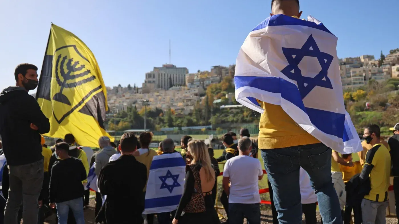 محتجون يحملون أعلام إسرائيلية ويظهرون في خلفية ملعب، يعبرون عن دعمهم لنادي بيتار القدس وسط جدل حول العنصرية في كرة القدم.