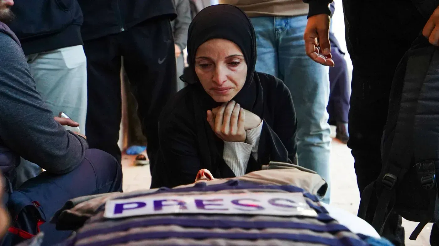 امرأة ترتدي الحجاب تجلس بجانب جثة صحفي فلسطيني، تحمل لافتة "PRESS"، تعبر عن الحزن وسط تجمع من الناس في غزة.