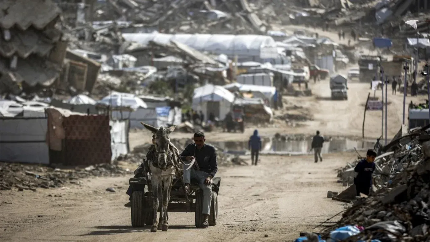 رجل يقود عربة تجرها حمار في منطقة مدمرة في غزة، حيث تظهر الخيام والمباني المدمرة نتيجة النزاع المستمر.