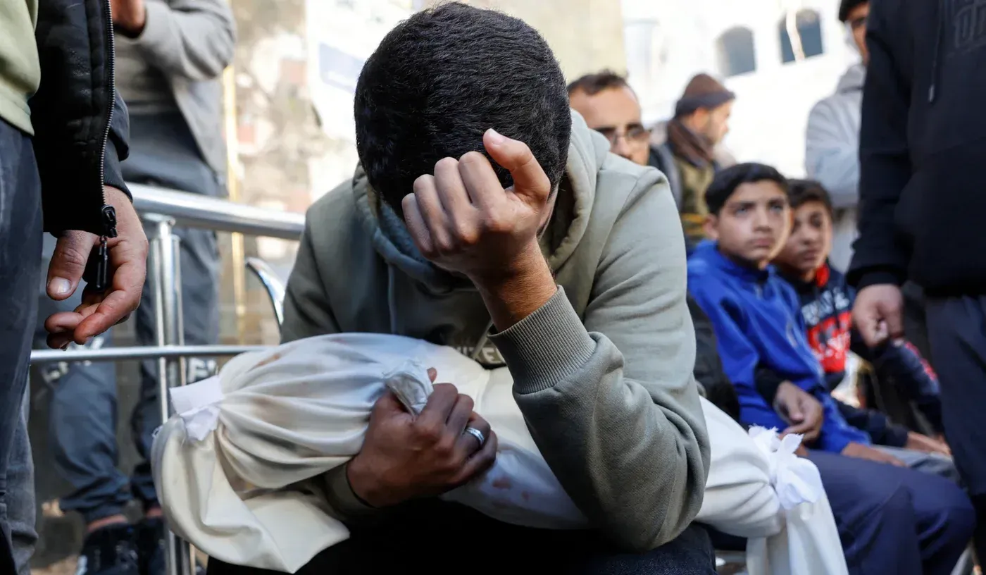 رجل يحمل طفلاً مغطىً بغطاء أبيض، يظهر عليه الحزن الشديد، بينما يتجمع آخرون في الخلف، في سياق مأساة قصف مدرسة في غزة.