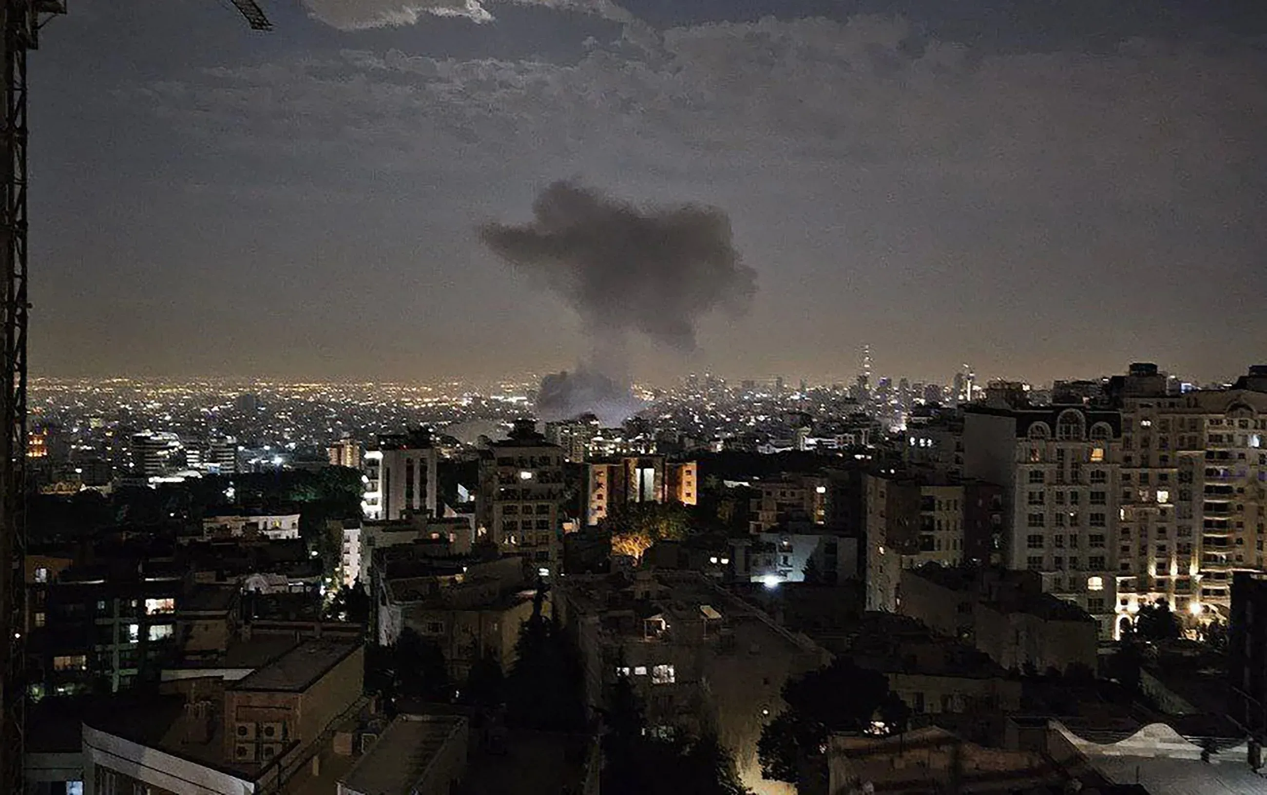 تصاعد دخان كثيف في سماء طهران بعد الهجمات الإسرائيلية على مواقع عسكرية، مع مشاهد للمدينة مضاءة ليلاً.