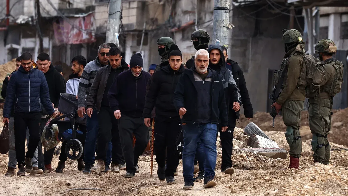 مشهد لعدد من الفلسطينيين يسيرون في منطقة متضررة، بينما تراقبهم قوات الاحتلال الإسرائيلي، مما يعكس تصاعد التوترات في الضفة الغربية.