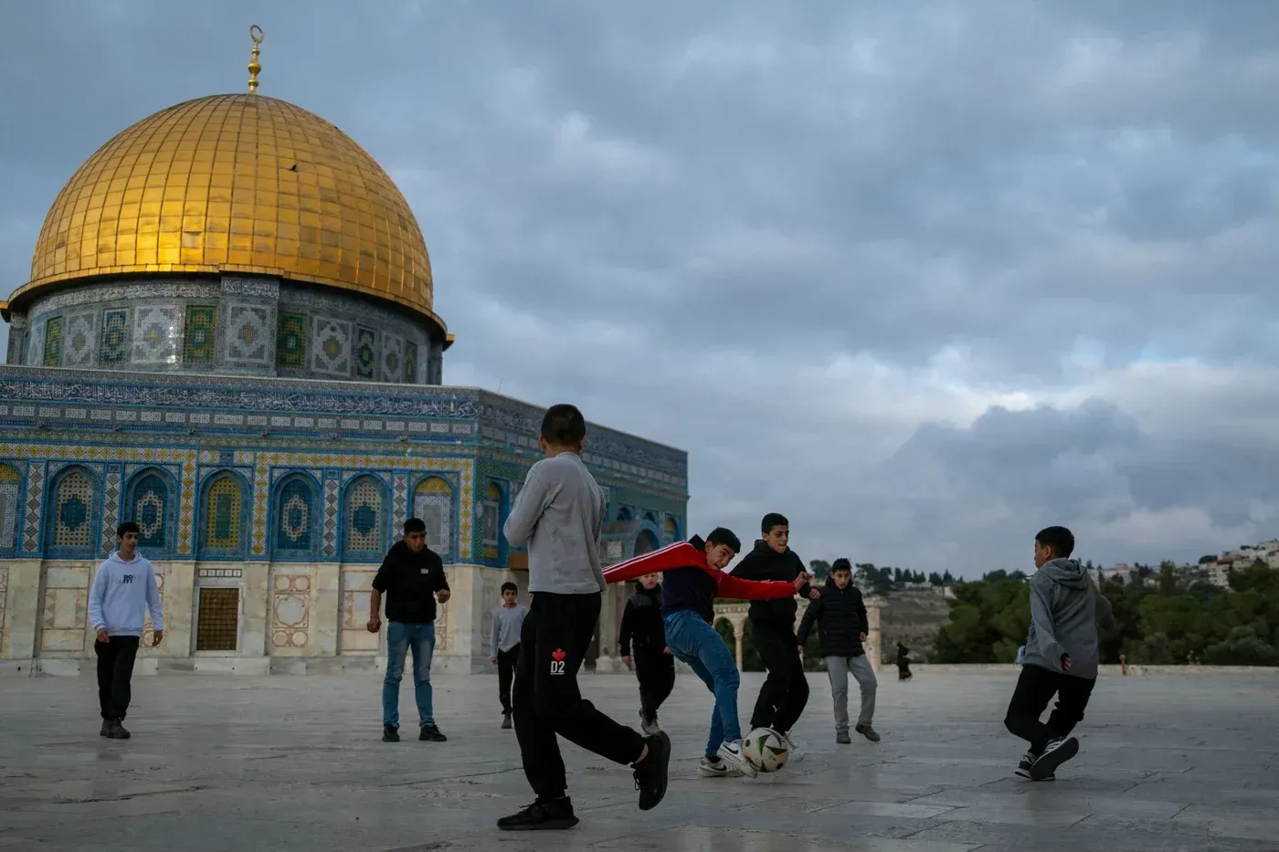 مجموعة من الأطفال يلعبون كرة القدم في ساحة المسجد الأقصى، مع قبة الذهب في الخلفية، في أجواء هادئة قبل رمضان.