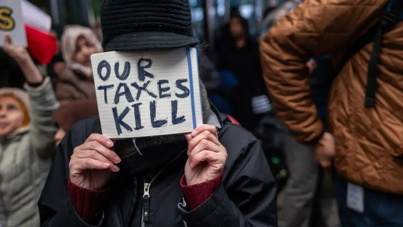 امرأة تحمل لافتة مكتوب عليها "ضرائبنا تقتل" خلال احتجاج، تعكس مشاعر المعارضة تجاه الدعم العسكري لإسرائيل.