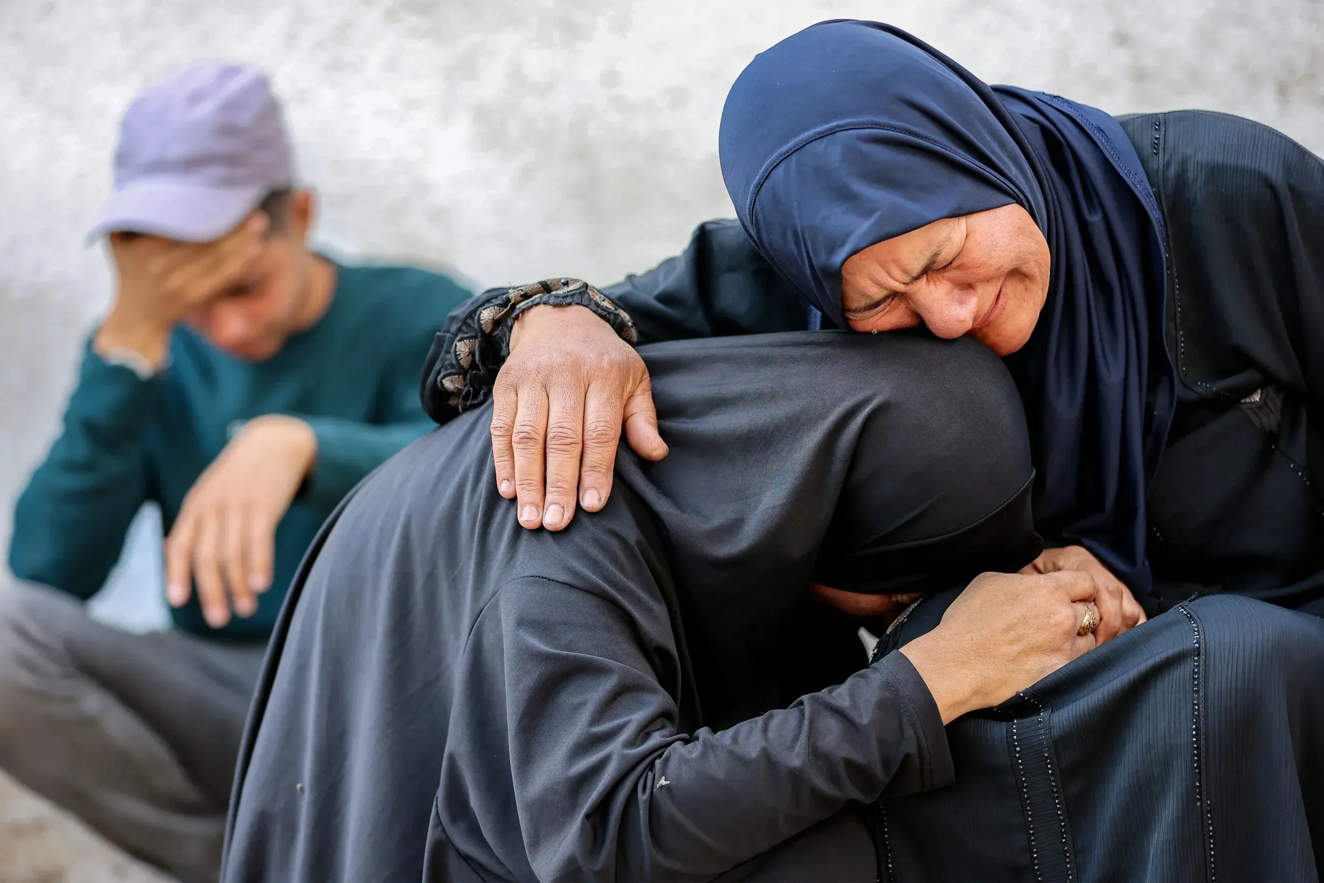 نساء فلسطينيات يعبرن عن حزنهن، بينما يظهر شاب في الخلفية. المشهد يعكس الألم والمعاناة الناتجة عن الهجمات الإسرائيلية في غزة.