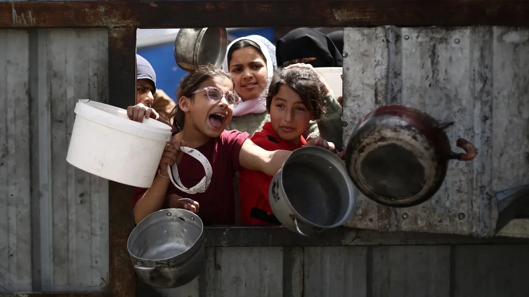 أطفال فلسطينيون يتجمعون حول نقطة توزيع، يحملون أواني للطعام، معبرين عن الحاجة الماسة للمساعدات الإنسانية في غزة.