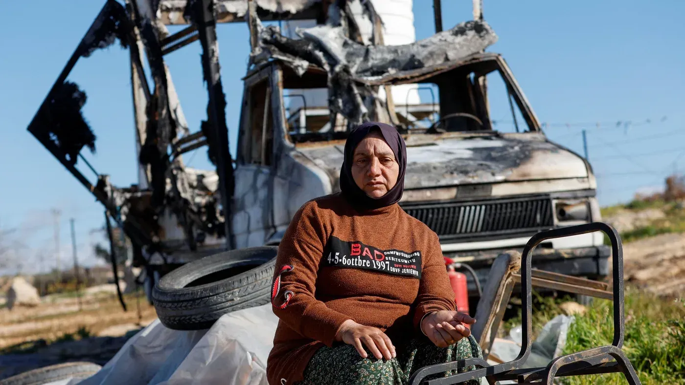 امرأة جالسة أمام شاحنة محترقة في منطقة متضررة، تعكس آثار الصراع في الأراضي الفلسطينية المحتلة وتسلط الضوء على معاناة المدنيين.