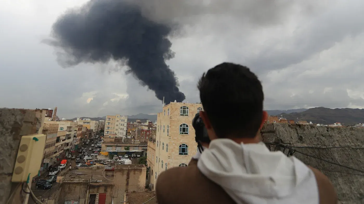 دخان أسود يتصاعد من موقع قصف في صنعاء، بينما يشاهد شخص من فوق سطح مبنى، مما يعكس آثار الغارات الإسرائيلية على المدينة.