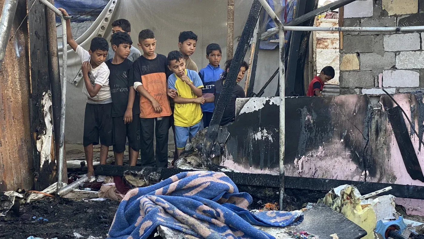 مجموعة من الأطفال يقفون بجوار خيمة محترقة في منطقة المواصي بخان يونس، تعبيرًا عن معاناتهم بعد الهجمات الإسرائيلية على النازحين.