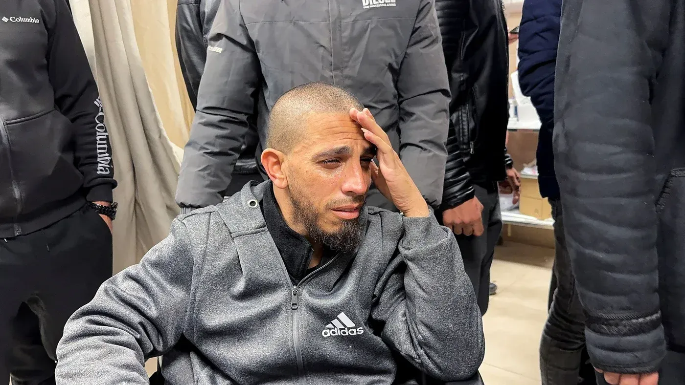 رجل يجلس في كرسي متحرك، يبدو عليه الحزن والقلق، محاطًا بأشخاص آخرين، في سياق الهجمات الإسرائيلية على جنين.