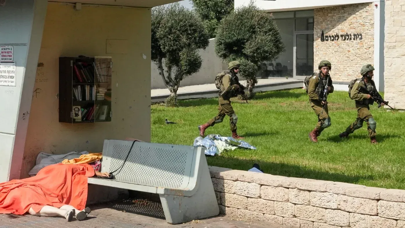 جنود إسرائيليون مسلحون يسيرون في منطقة عامة بعد هجوم حماس، مع وجود شخص ممدد على مقعد بجوارهم، مما يعكس حالة الفوضى والذعر.