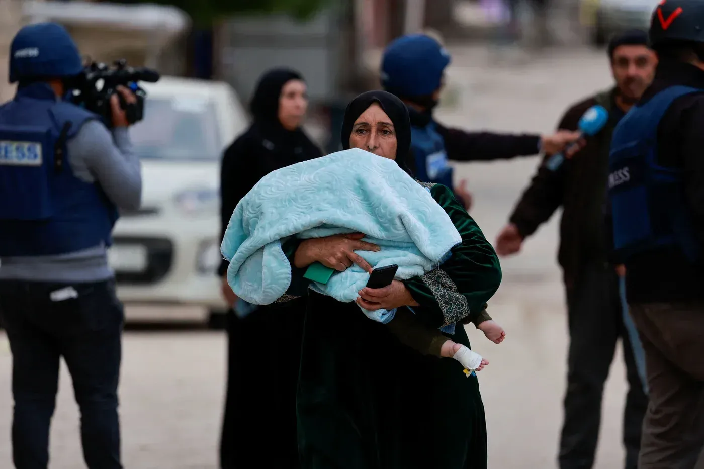 امرأة تحمل طفلاً مغطى ببطانية، بينما تظهر حشود من الصحفيين في خلفية مشهد متوتر في جنين وسط التوترات الحادة.