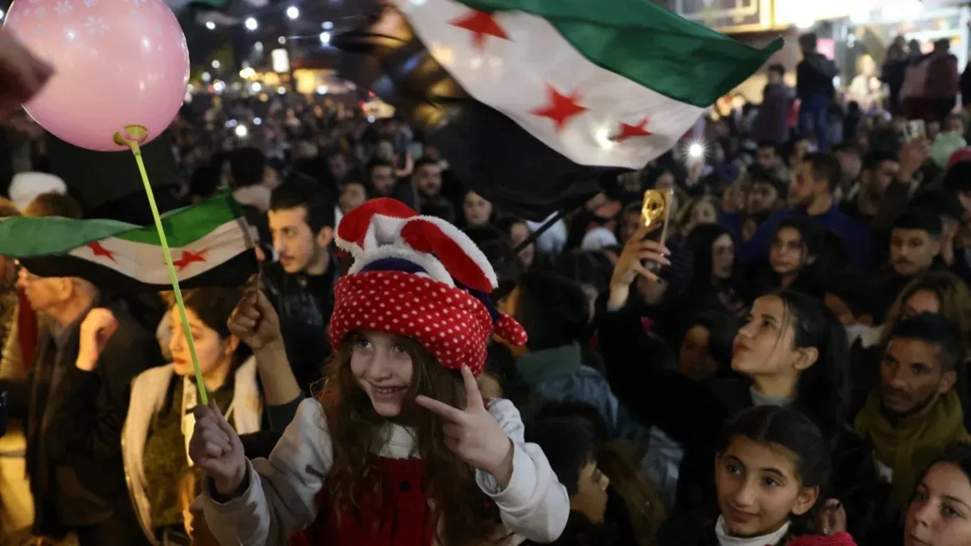 طفلة ترتدي قبعة حمراء مزخرفة تحمل بالونًا ورديًا، وسط حشد من الناس يحملون الأعلام السورية، في أجواء احتفالية تعكس التوترات الحالية في سوريا.