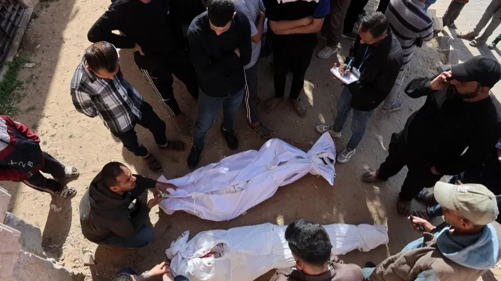 تجمع حشد من الناس حول جثتين مغطاتين في موقع الهجوم الإسرائيلي على متطوعين وصحفيين في غزة، مما يعكس حجم المأساة الإنسانية.