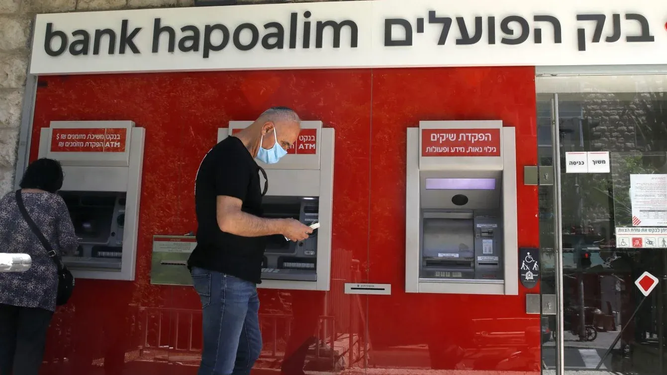 رجل يرتدي كمامة يستخدم صرافاً آلياً أمام فرع بنك Hapoalim الإسرائيلي وسط تصاعد التوترات المالية وتأثيرات وقف خدمات المراسلة المصرفية على الاقتصاد الفلسطيني.