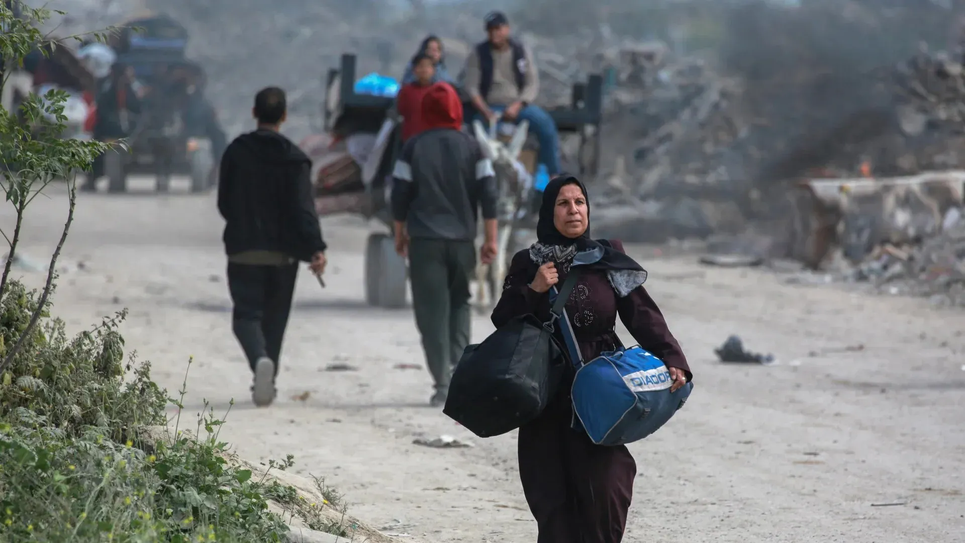 امرأة فلسطينية تحمل حقائب وتبدو في حالة من التوتر أثناء سيرها في منطقة مدمرة، وسط مجموعة من الأشخاص في الخلفية.
