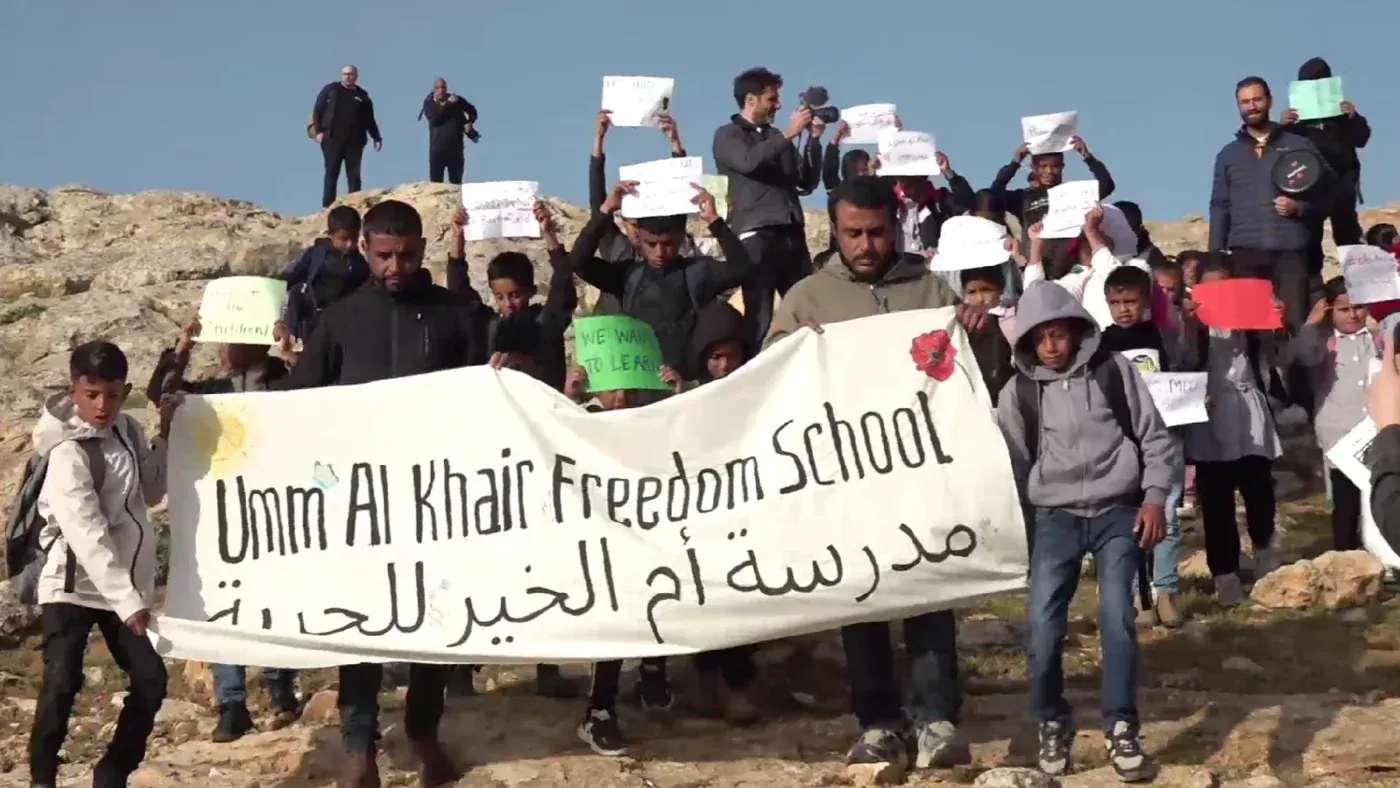 طلاب فلسطينيون في قرية أمّ الخير يحملون لافتات خلال احتجاج للمطالبة بحقهم في التعليم، وسط انتشار قوات الاحتلال الإسرائيلي.