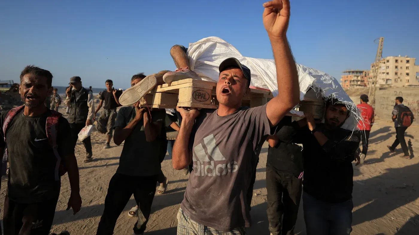 محتجون يحملون جثمانًا ملفوفًا في قماش أبيض، يعبرون عن حزنهم في منطقة تعرضت للقصف في غزة، وسط أجواء من الفوضى والدمار.