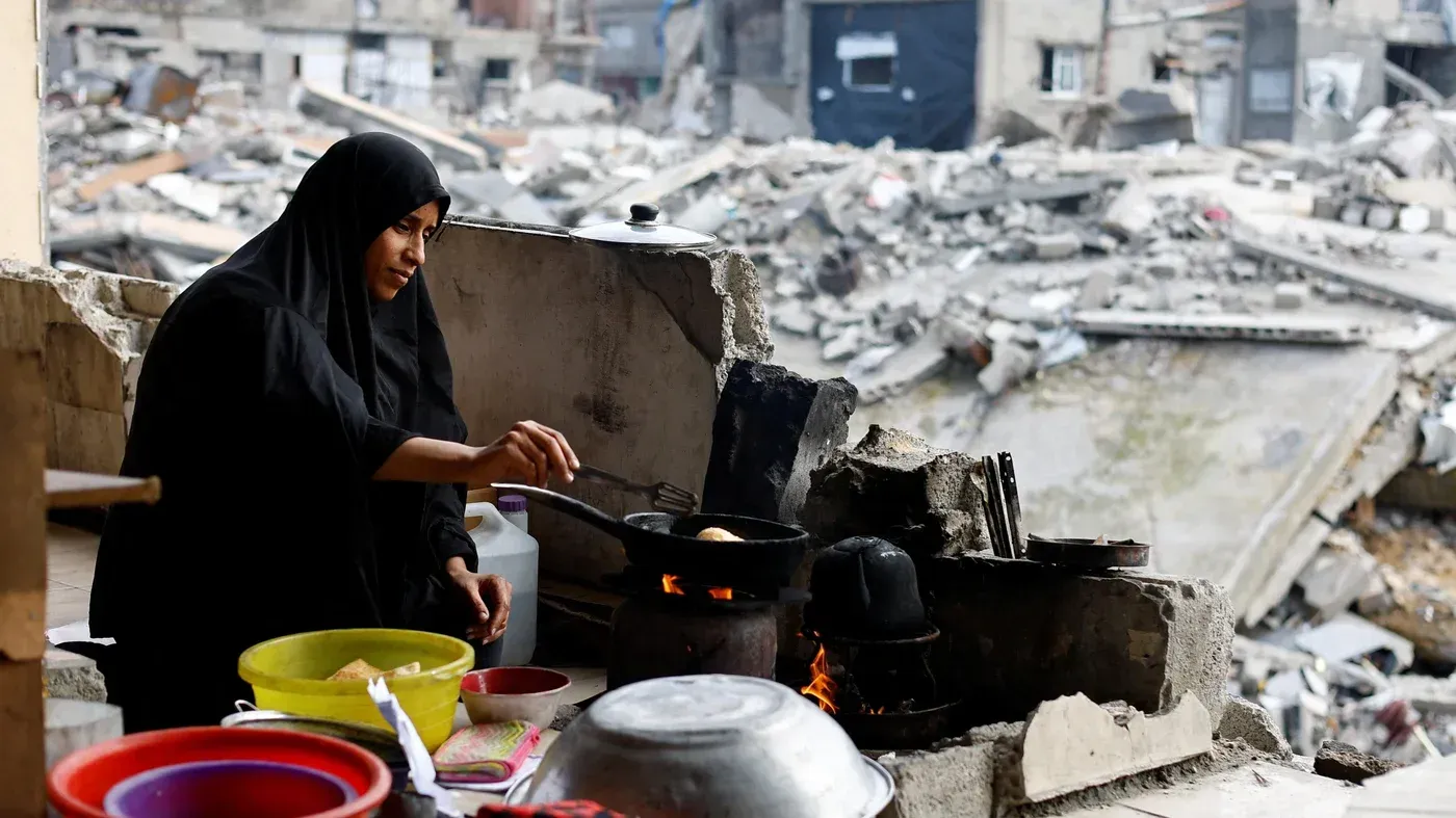 امرأة ترتدي الحجاب تطبخ في مطبخ بدائي وسط أنقاض مبانٍ مدمرة في غزة، تعكس معاناة السكان في ظل الظروف الصعبة.