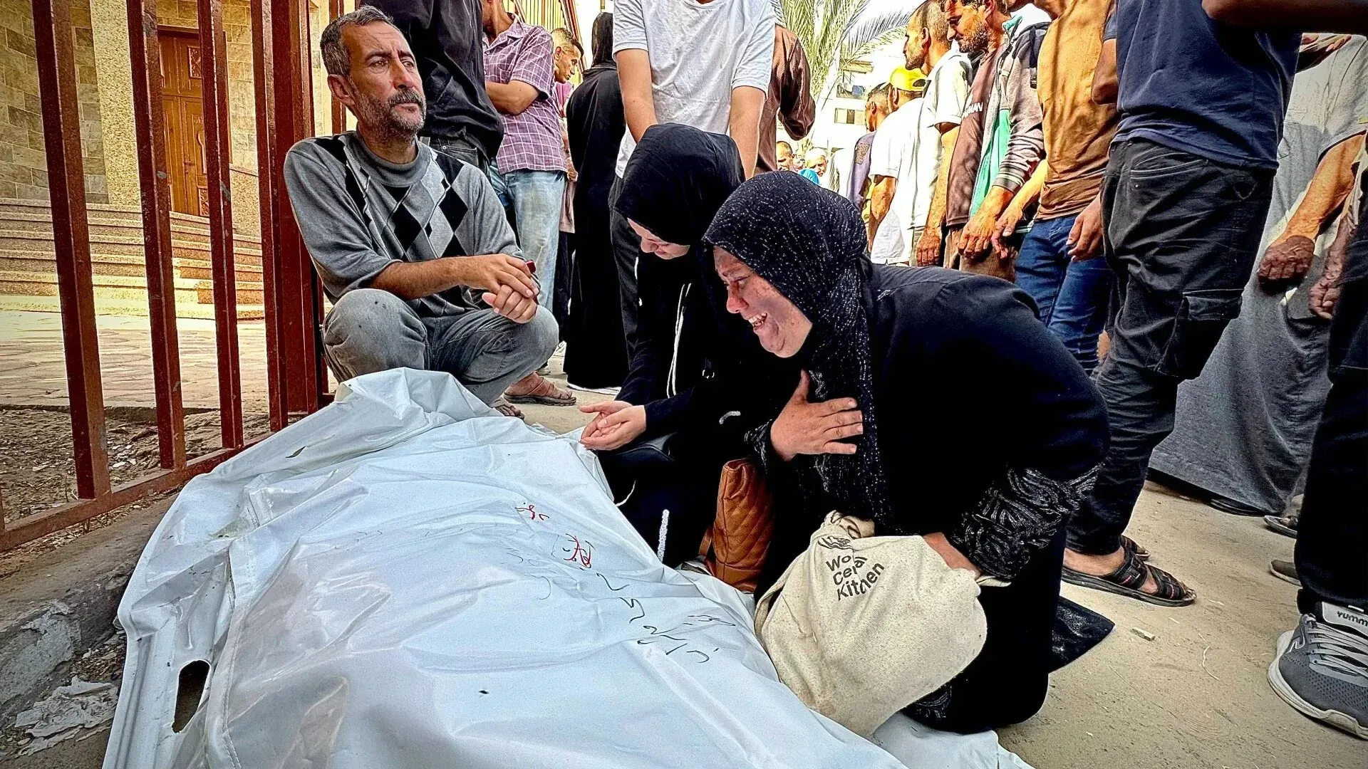 مشهد مؤلم لعائلة فلسطينية تتجمع حول جثة مغطاة، مع تعبيرات الحزن والصدمة، في سياق العنف المستمر في غزة.