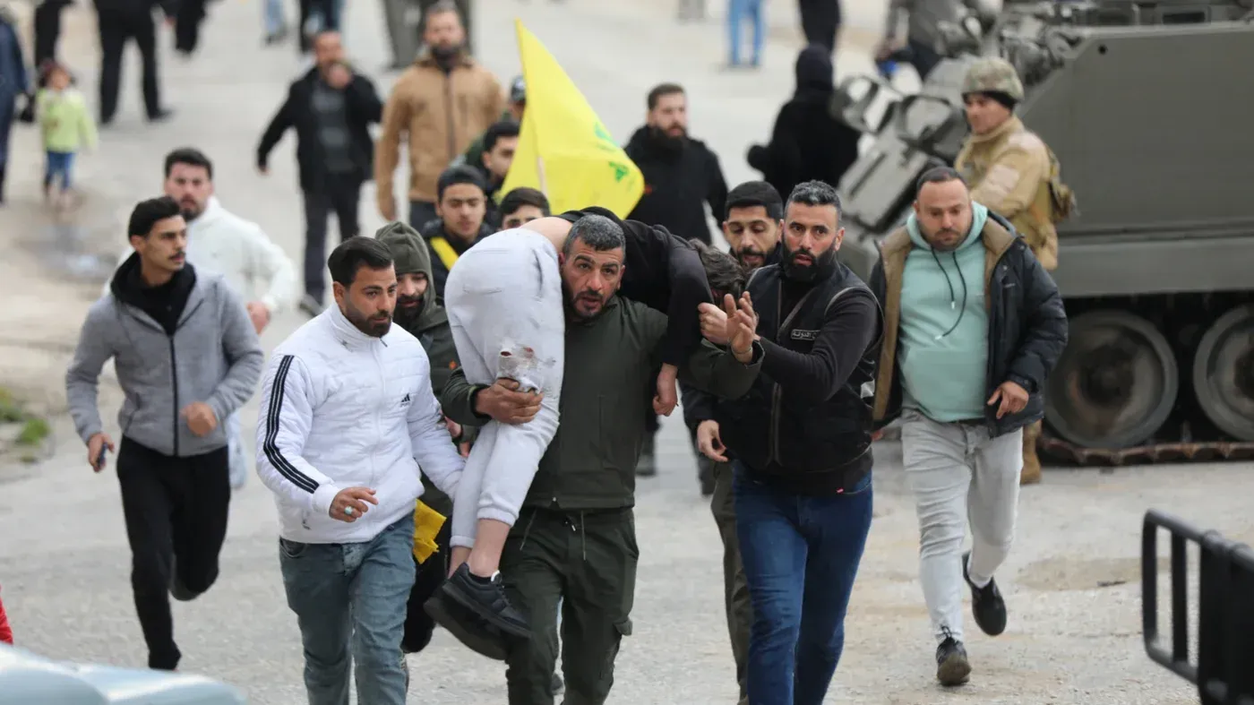 مجموعة من اللبنانيين يحملون شخصًا مصابًا بينما يتجهون نحو قوات الاحتلال الإسرائيلي، مع وجود علم لحزب الله في الخلفية.