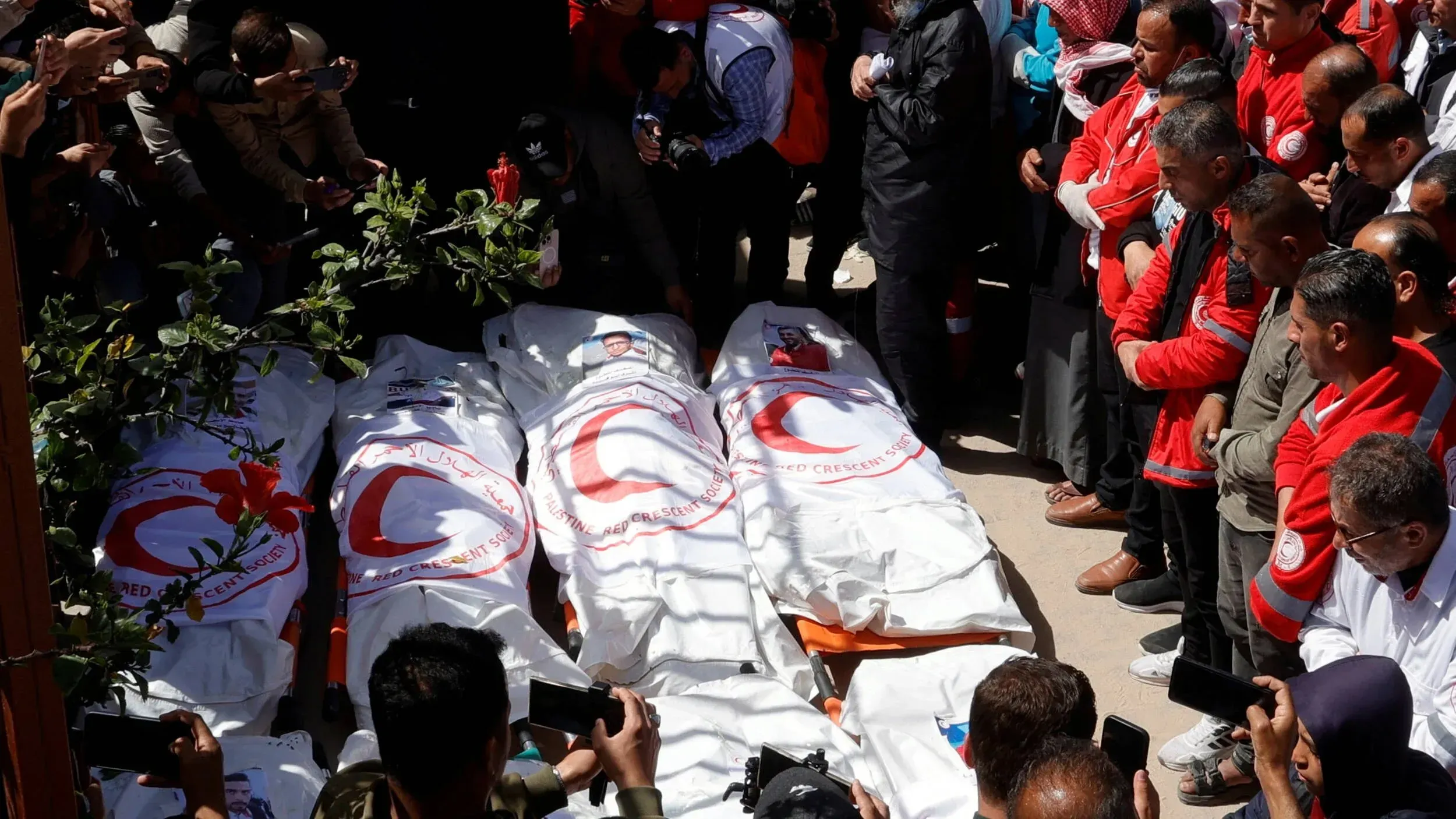 تجمع حشود من الناس في جنازة لعمال الطوارئ الفلسطينيين الذين قُتلوا، مع وجود جثث مغطاة بأعلام الهلال الأحمر، تعبيرًا عن الحزن.