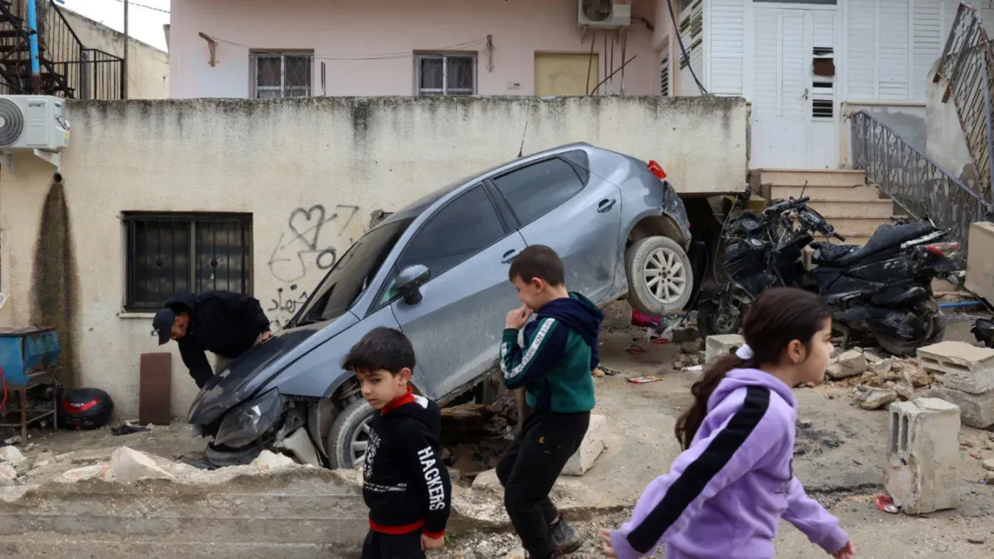 أطفال فلسطينيون يلعبون في شارع مدمّر، حيث يظهر سيارة مقلوبة وأخرى متضررة، مما يعكس آثار الهجمات الإسرائيلية على الضفة الغربية.