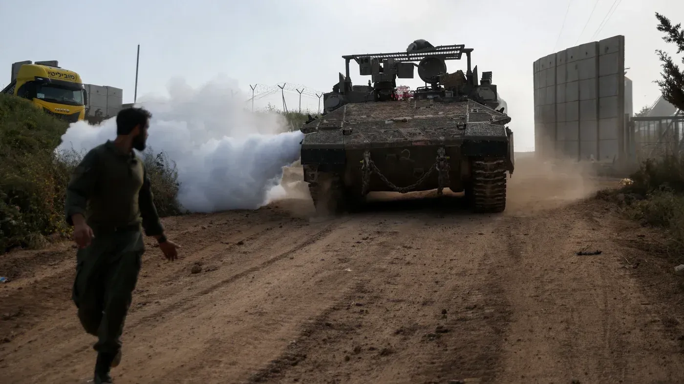دبابة إسرائيلية تطلق دخاناً على طريق ترابي في جنوب لبنان، بينما يركض جندي نحوها، مما يعكس تصاعد التوترات العسكرية في المنطقة.