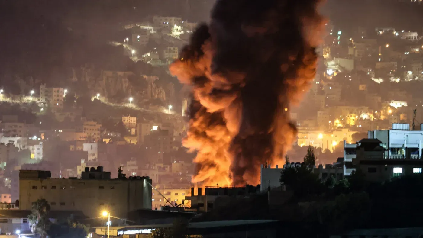 تصاعد الدخان من انفجار في منطقة نابلس، مع إضاءة المدينة في الخلفية، مما يعكس تصاعد التوترات والاعتداءات في الضفة الغربية.