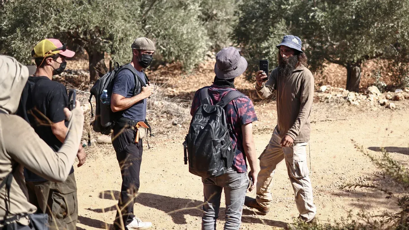 مجموعة من المستوطنين الإسرائيليين في الضفة الغربية، يتحدثون معًا في منطقة زراعية، مع وجود أشجار الزيتون في الخلفية.