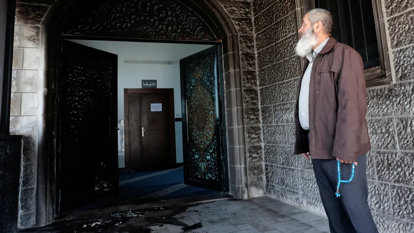 رجل يقف أمام مدخل مسجد أبو بكر الصديق المحترق في قرية تل، حيث تظهر آثار الحريق على الأبواب والجدران.