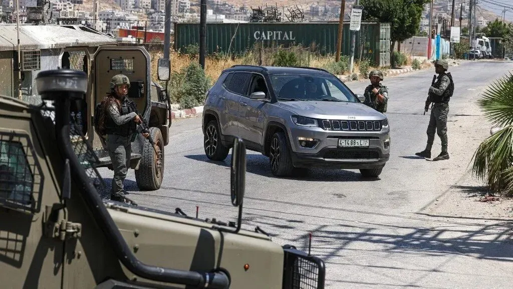 جنود إسرائيليون يقفون عند نقطة تفتيش في الضفة الغربية، مع وجود مركبة مدنية. تعكس الصورة التوترات الأمنية المستمرة في المنطقة.