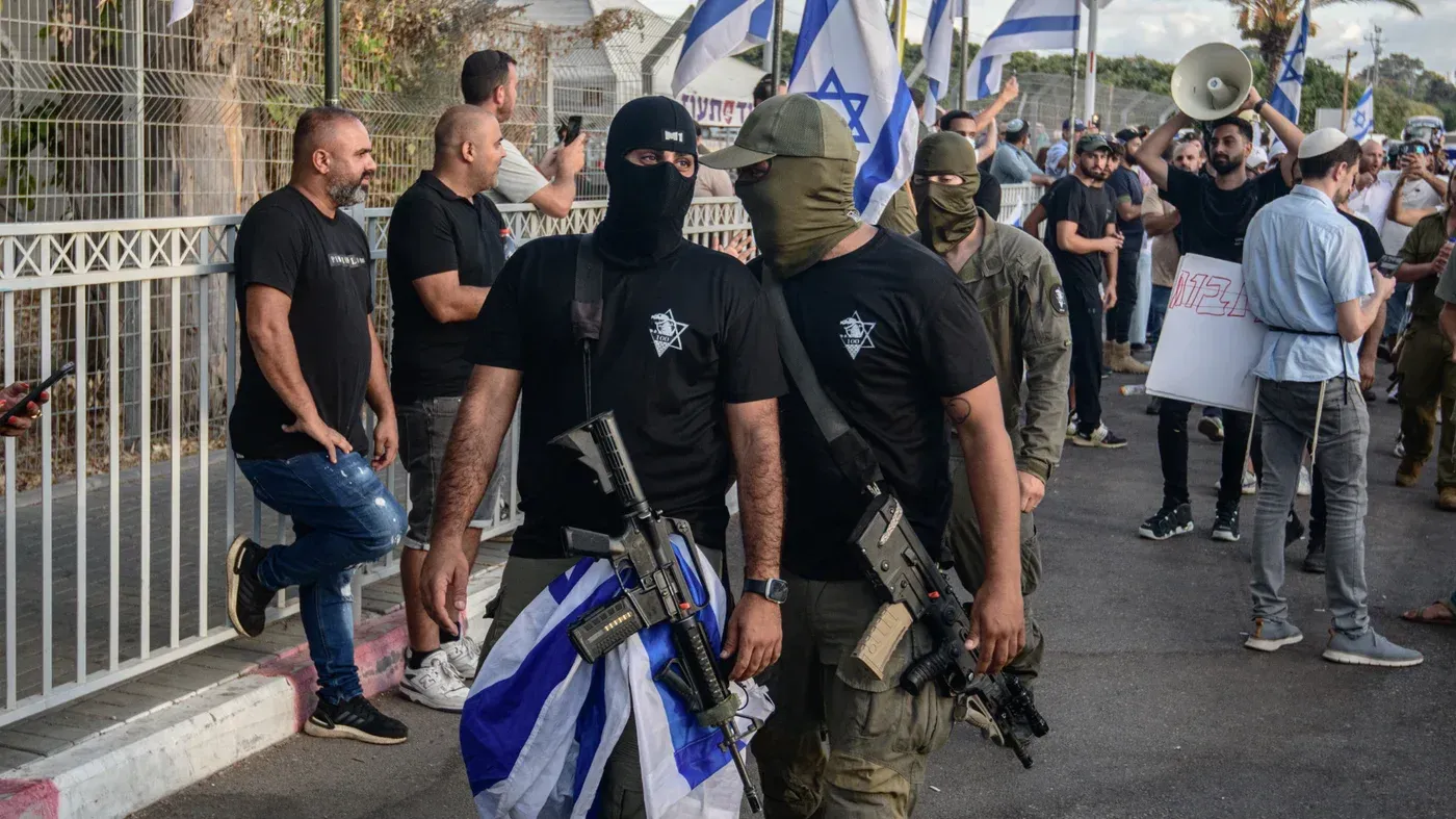 محتجون يرتدون أقنعة سوداء ويحملون أسلحة، يتظاهرون في شارع مع أعلام إسرائيلية، وسط أجواء متوترة تعكس الانتهاكات بحق الفلسطينيين.