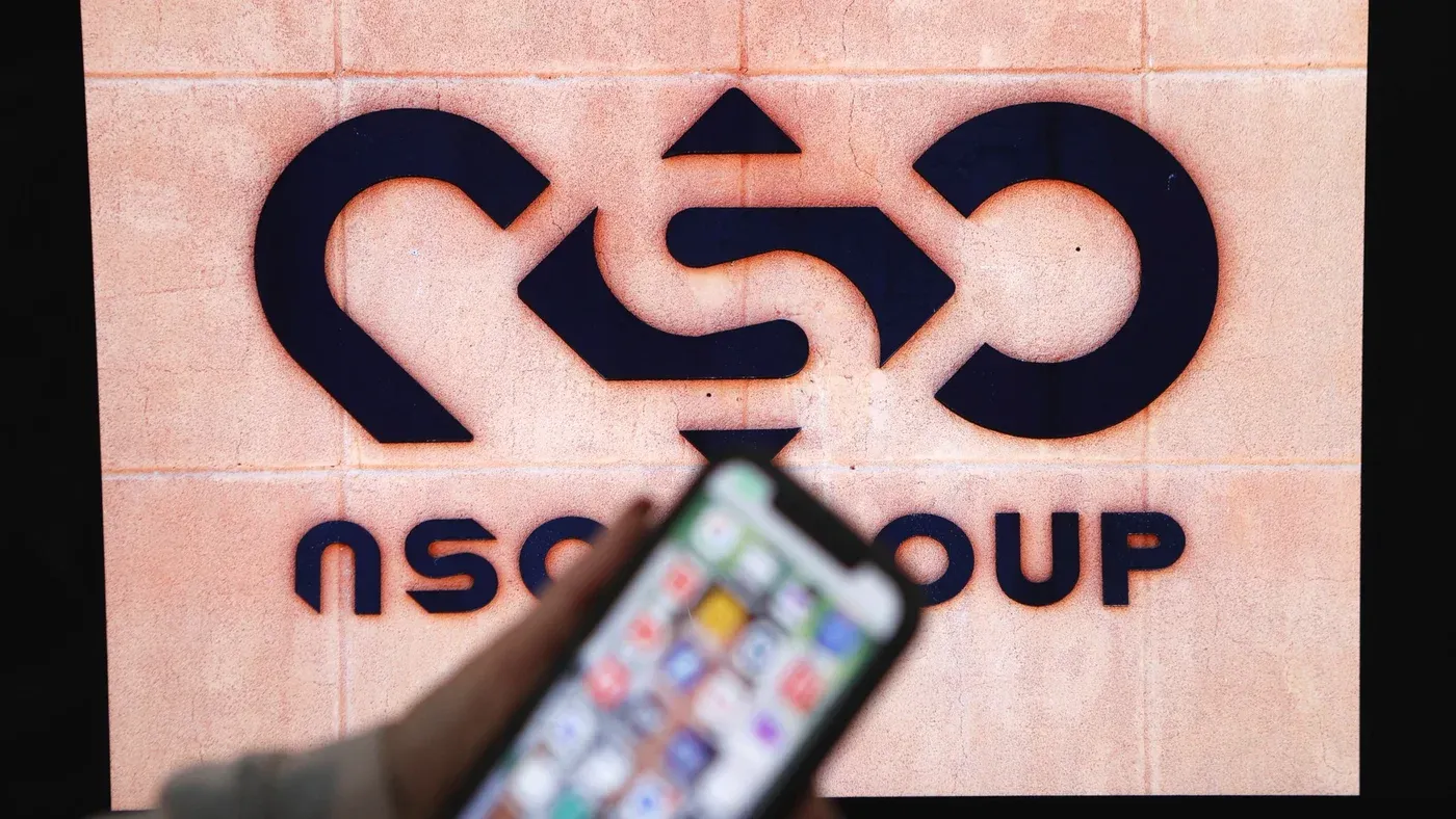 شعار مجموعة NSO يظهر على جدار، مع هاتف ذكي في المقدمة، مما يبرز محاولات استهداف مستخدمي WhatsApp عبر روابط خبيثة.