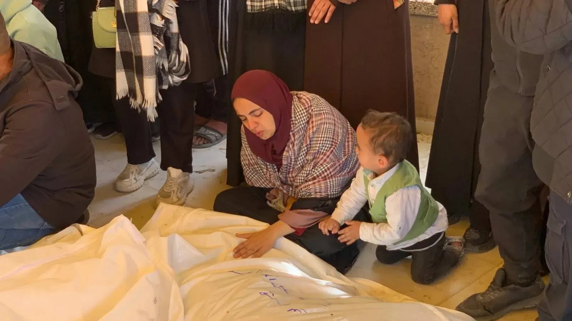 امرأة ترتدي حجابًا تجلس بجانب طفل صغير، وتظهر علامات الحزن على وجهها، بينما يحيط بهم أشخاص آخرون في موقع جنازة، مع وجود جثث مغطاة بأقمشة بيضاء.