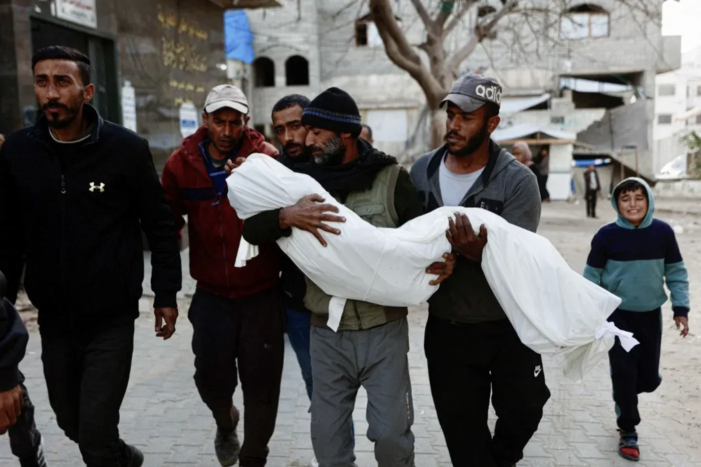 رجال يحملون جثمان طفل مغطى ببطانية بيضاء في شوارع غزة، مع وجود طفل صغير يبتسم في الخلفية، في سياق الغارات الإسرائيلية.