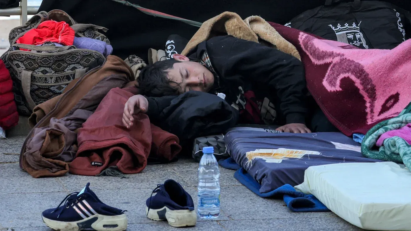 طفل نائم على الأرض بين الأغطية والحقائب، يعكس معاناة الأطفال النازحين في لبنان جراء الحرب الإسرائيلية وتأثيرها المدمر على حياتهم.