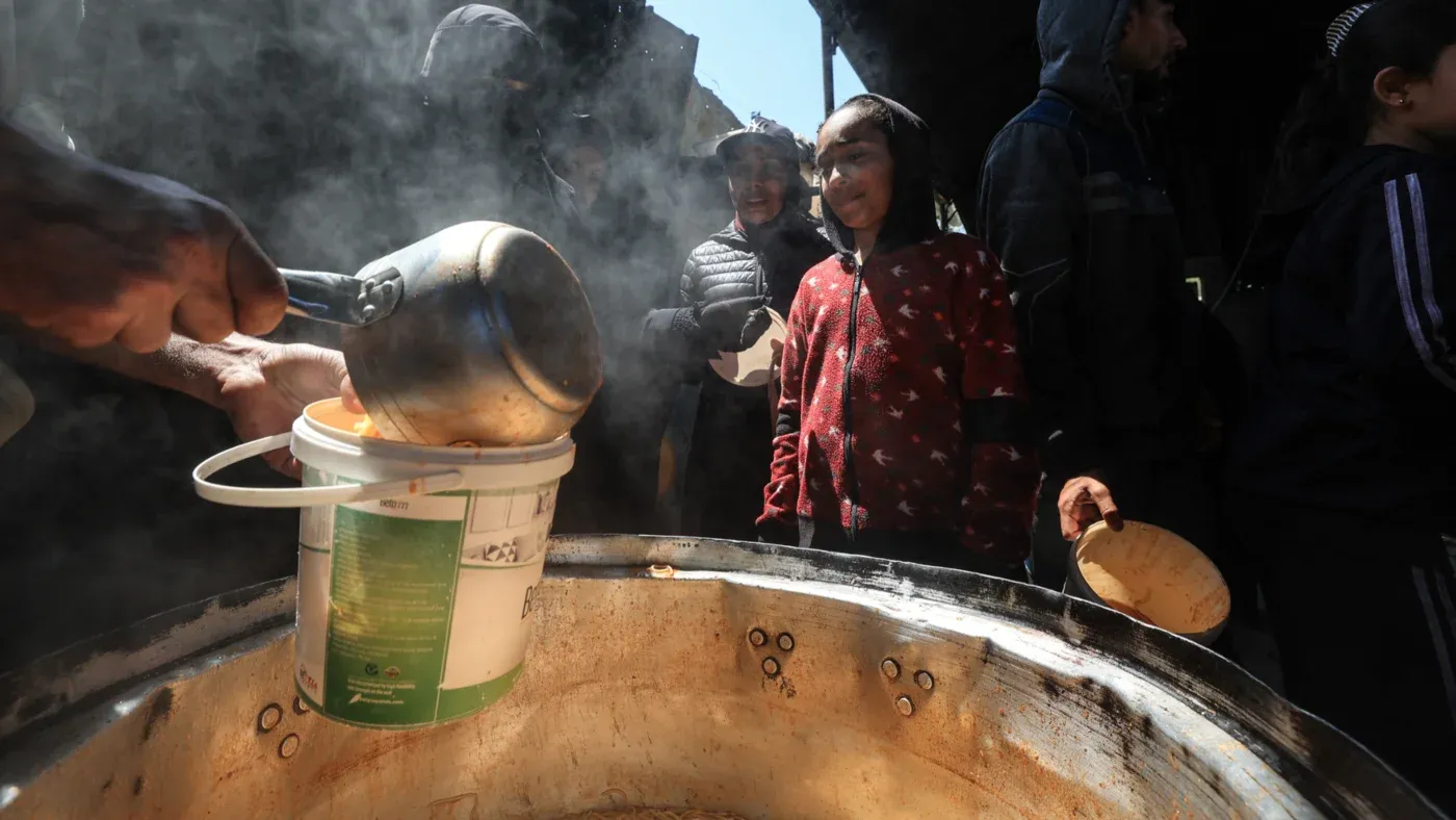 تظهر الصورة شخصًا يوزع الطعام من وعاء كبير على مجموعة من الأطفال والبالغين في غزة، وسط أجواء من الضباب والدخان، مما يعكس أزمة التجويع المستمرة.