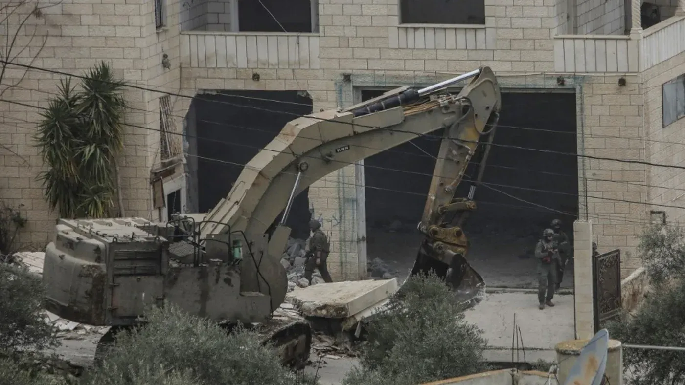 جرافة تعمل على هدم مبنى في الضفة الغربية المحتلة، بينما يتواجد جنود إسرائيليون في المنطقة، مما يعكس تصعيد عمليات التهجير والمصادرة.
