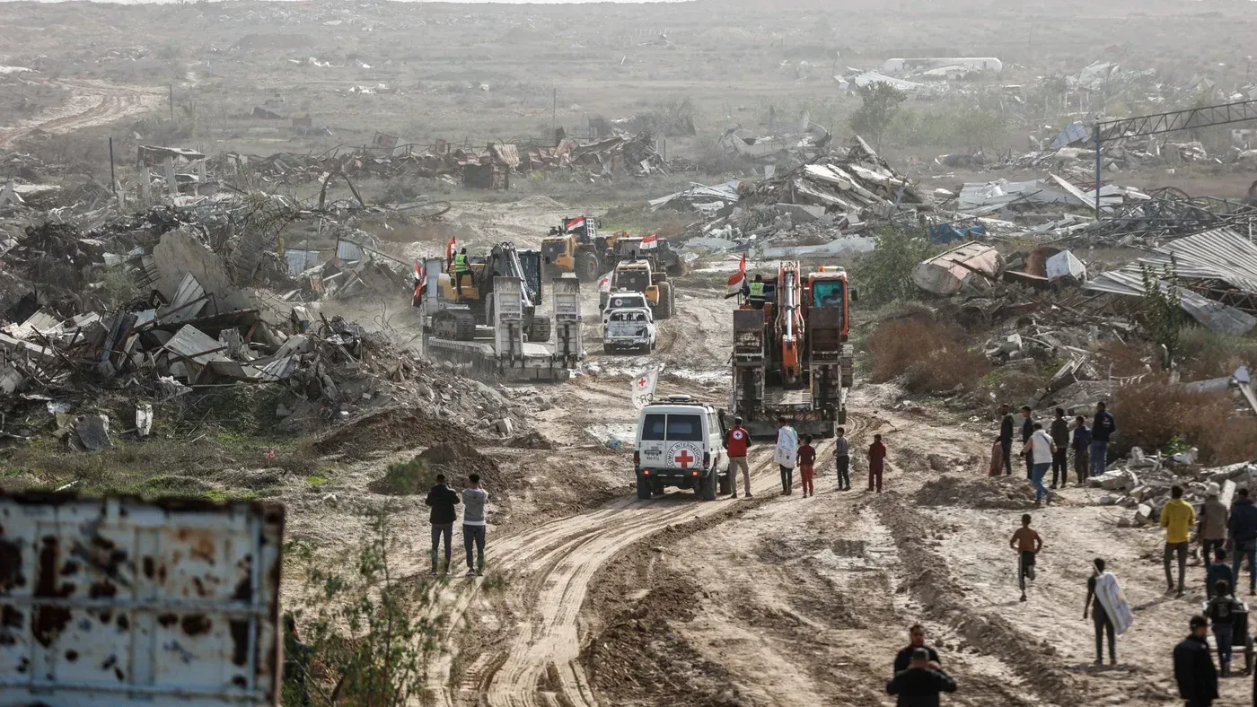 تظهر الصورة مشهدًا من منطقة مدمرة، حيث تتواجد آليات ثقيلة وسيارات إسعاف، مع أشخاص يتجولون في الأرجاء. تعكس الصورة آثار النزاع وتأثيره على المدنيين.