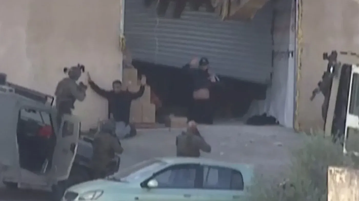 مواجهات في جنين تظهر فلسطينيين غير مسلحين يرفعان أيديهما، بينما تتواجد قوات الاحتلال الإسرائيلية في الخلفية.