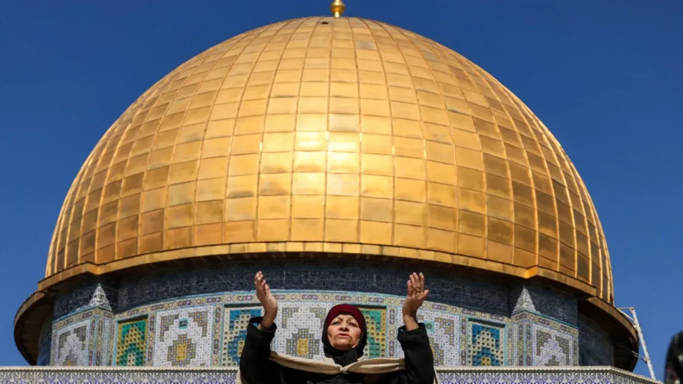 امرأة تؤدي الصلاة أمام قبة المسجد الأقصى، مع خلفية معمارية مميزة، في ظل تصاعد التوترات خلال شهر رمضان.