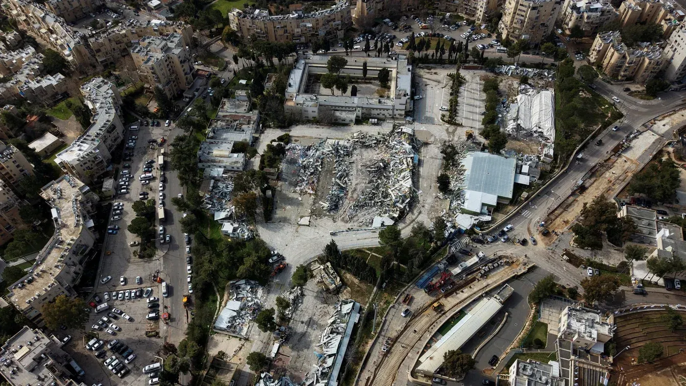 تظهر الصورة مشهدًا جويًا لمجمع الأونروا المدمر في القدس الشرقية، حيث تبرز الأنقاض وسط المباني السكنية والسيارات.